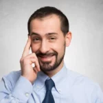 Homme souriant en chemise et cravate, posant un doigt sur sa tempe en signe d’astuce ou de réflexion.