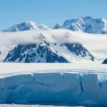 Panorama de l’Antarctique avec un glacier bleu en premier plan, des montagnes enneigées en arrière-plan et une nappe de nuages bas flottant entre les sommets.