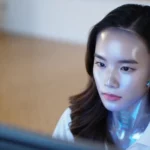 Femme robot humanoïde au visage métallique travaillant devant un écran d’ordinateur.