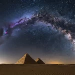 Photographie nocturne spectaculaire de la Voie lactée s’étendant en arc de cercle au-dessus des pyramides de Gizeh, en Égypte, sous un ciel étoilé cristallin, avec des dunes de sable au premier plan.