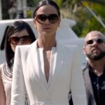 Femme en costume blanc et lunettes de soleil marchant devant une équipe de gardes du corps en noir