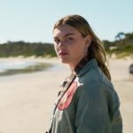 Une jeune femme sérieuse regarde vers la caméra sur une plage, avec un appareil photo autour du cou — scène de la série The Survivors sur Netflix