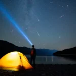 Personne observant une pluie d’étoiles filantes sous un ciel dégagé, avec une tente éclairée au bord d’un lac