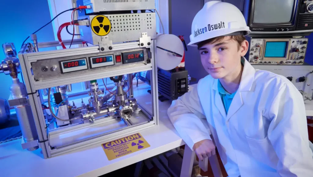 Jackson Oswalt, adolescent en blouse de laboratoire, pose à côté de son réacteur de fusion nucléaire artisanal.