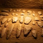 Outils préhistoriques en pierre taillée découverts lors d’une fouille archéologique, éclairés dans une cavité de terre.