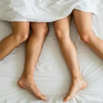 Deux personnes allongées côte à côte sous une couverture blanche, symbolisant l’intimité et les relations sexuelles.