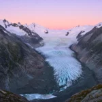 Vue d’un glacier alpin descendant entre deux chaînes de montagnes, éclairé par une lumière rosée au lever du soleil.