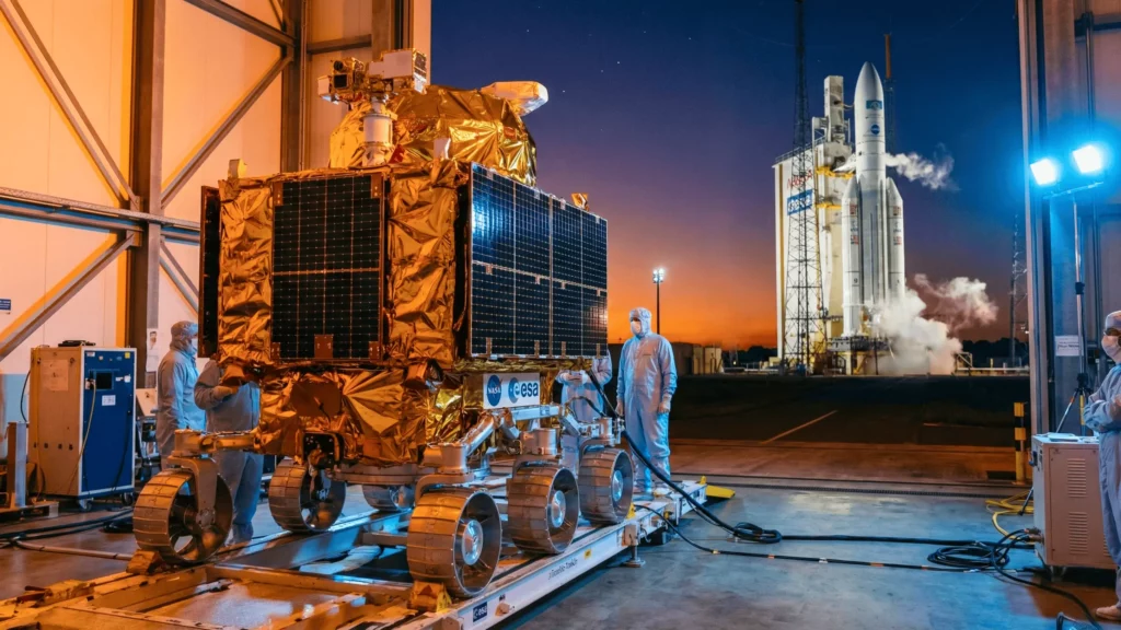 Rover de l’ESA en préparation dans un hangar avec des ingénieurs, et fusée Ariane prête au lancement au coucher du soleil