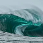 Vague géante turquoise en pleine mer, se courbant avec force et projetant une écume blanche sous un ciel gris.