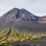 Volcan aux pentes de cendres noires avec un cratère ouvert, strates colorées visibles sur les parois, dans un paysage du Kamtchatka en Russie.