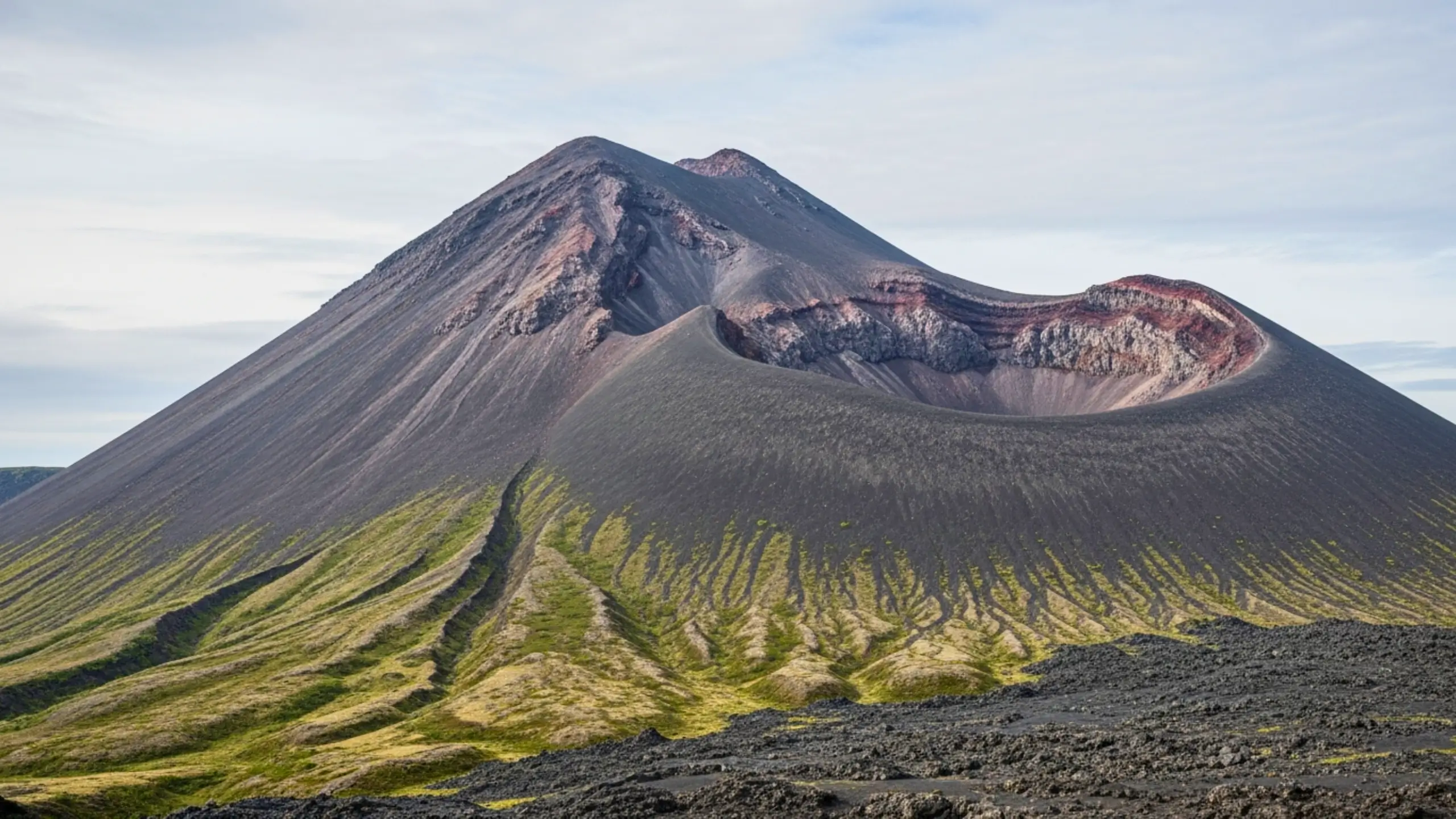 Volcan aux pentes de cendres noires avec un cratère ouvert, strates colorées visibles sur les parois, dans un paysage du Kamtchatka en Russie.