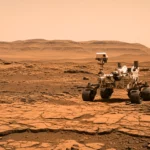 Rover martien Perseverance sur une plaine rocheuse de Mars, avec collines à l’horizon sous un ciel orangé.