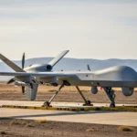Drone militaire de type UAV posé sur une piste, avec train d’atterrissage sorti, dans un paysage désertique avec montagnes au loin.