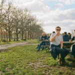 Personnes assises à des tables de pique-nique en extérieur, buvant et discutant sous un soleil d’hiver, avec des arbres nus et un chemin boueux à proximité.