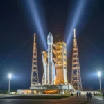 Fusée Ariane éclairée par des projecteurs sur son pas de tir de nuit, entourée de structures métalliques et de personnel au sol.