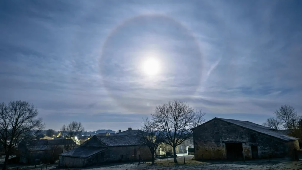 Halo circulaire autour de la Lune dans un ciel voilé, au-dessus de maisons et d’arbres d’un village rural en Charente.