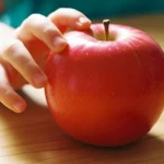 Main d'un jeune enfant s'approchant d'une pomme rouge fraîche posée sur une table en bois clair sous une lumière naturelle.
