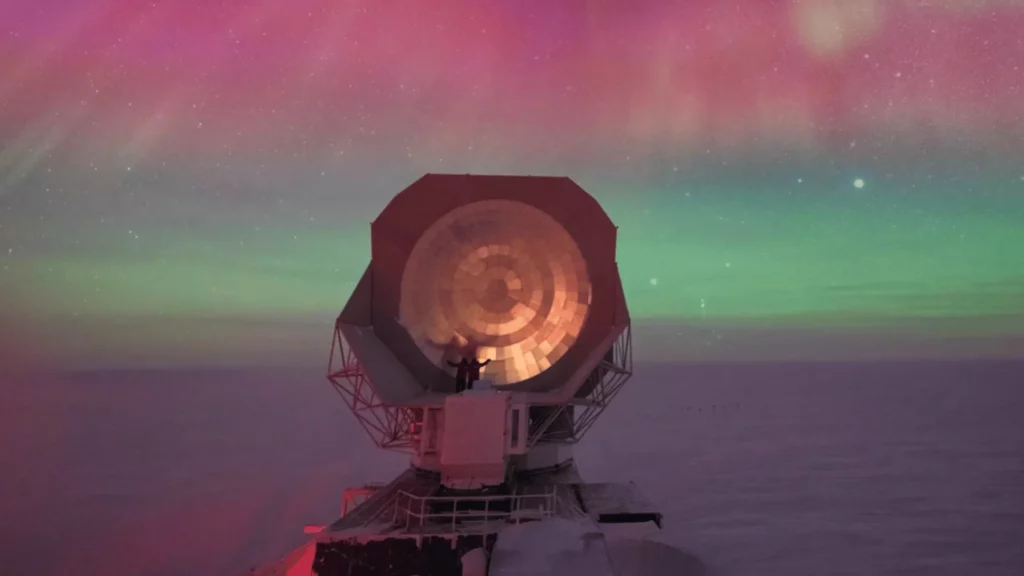 Un télescope géant pointé vers le ciel antarctique sous une aurore boréale multicolore, avec une silhouette humaine à sa base.