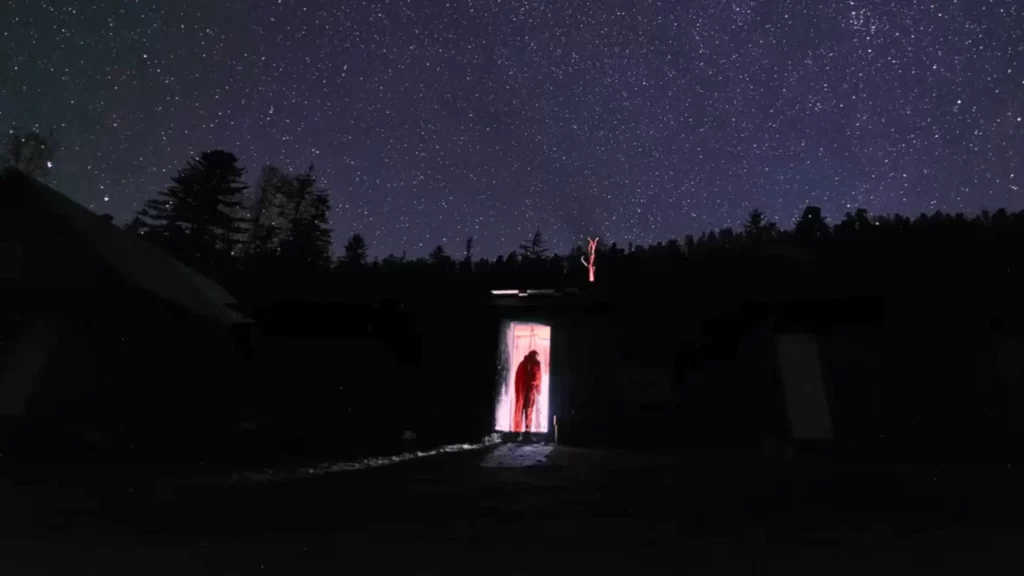 Une silhouette humaine se tient dans l’encadrement lumineux d’une porte ouverte, sous un ciel nocturne densément étoilé.