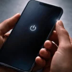 Une main tient un smartphone en train de redémarrer, écran sombre avec l’icône d’alimentation au centre.