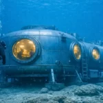 Un plongeur s’approche d’un habitat humain en acier installé à 200 mètres de profondeur, avec des chercheurs visibles à l’intérieur à travers des hublots éclairés.
