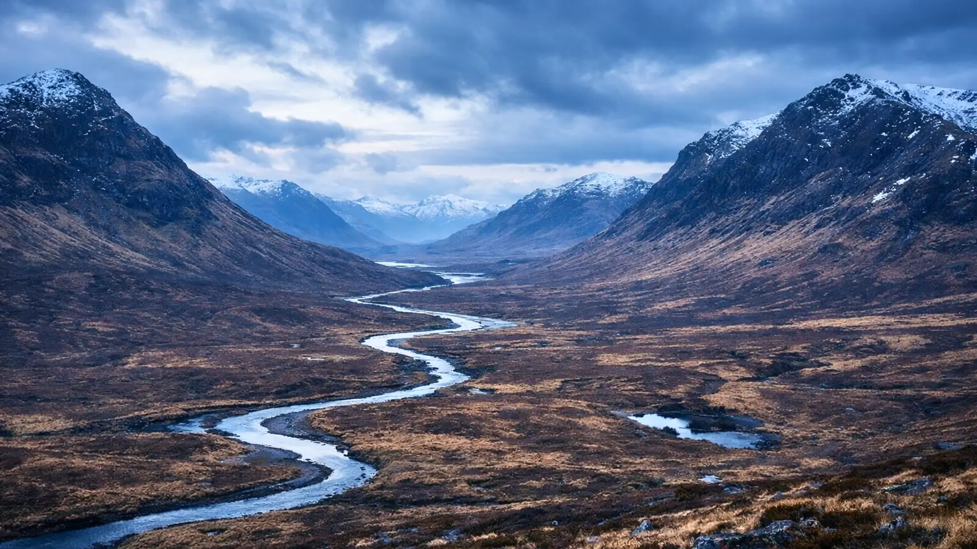 Vue panoramique des Highlands écossais avec une vallée sauvage et une rivière sinueuse sous un ciel nuageux dramatique.