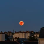 Lune de Sang rougeoyant au-dessus des toits d’une ville au crépuscule lors d’une éclipse lunaire totale