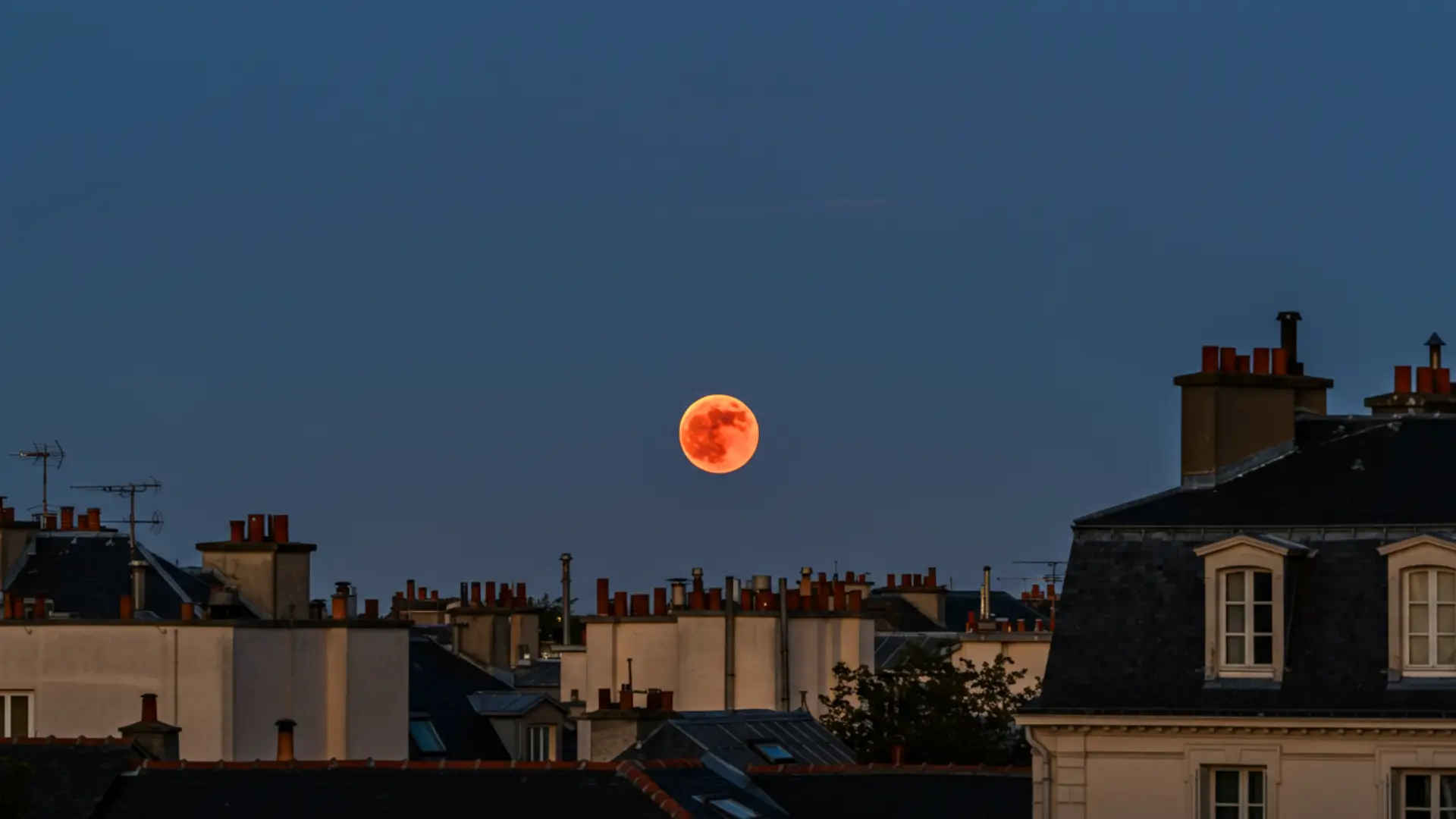 Lune de Sang rougeoyant au-dessus des toits d’une ville au crépuscule lors d’une éclipse lunaire totale