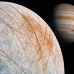 Vue réaliste de la lune Europe montrant sa surface glacée striée de fractures, avec Jupiter en arrière-plan dans l’espace.