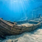 Une ancienne barge cérémonielle repose sous l’eau dans le port englouti d’Alexandrie, partiellement enfouie dans le sable, entourée de colonnes en ruine.