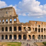Le Colisée de Rome apparaît en vue large sous un ciel bleu lumineux, avec quelques touristes à sa base et ses arches nettement visibles.