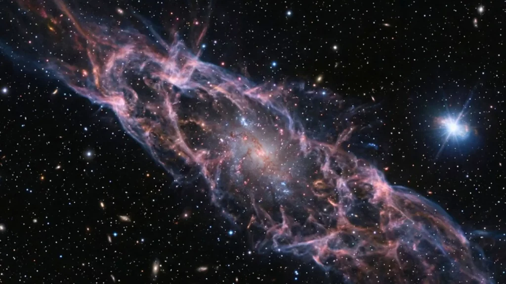 Une minuscule galaxie naine apparaît dans un vaste courant de gaz intergalactique, avec la trace lumineuse d’une collision d’étoiles à neutrons dans l’espace profond.