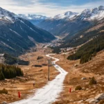 Une piste de ski étroite couverte d’un mince ruban de neige artificielle traverse une montagne alpine presque sans neige, avec des télésièges immobiles.