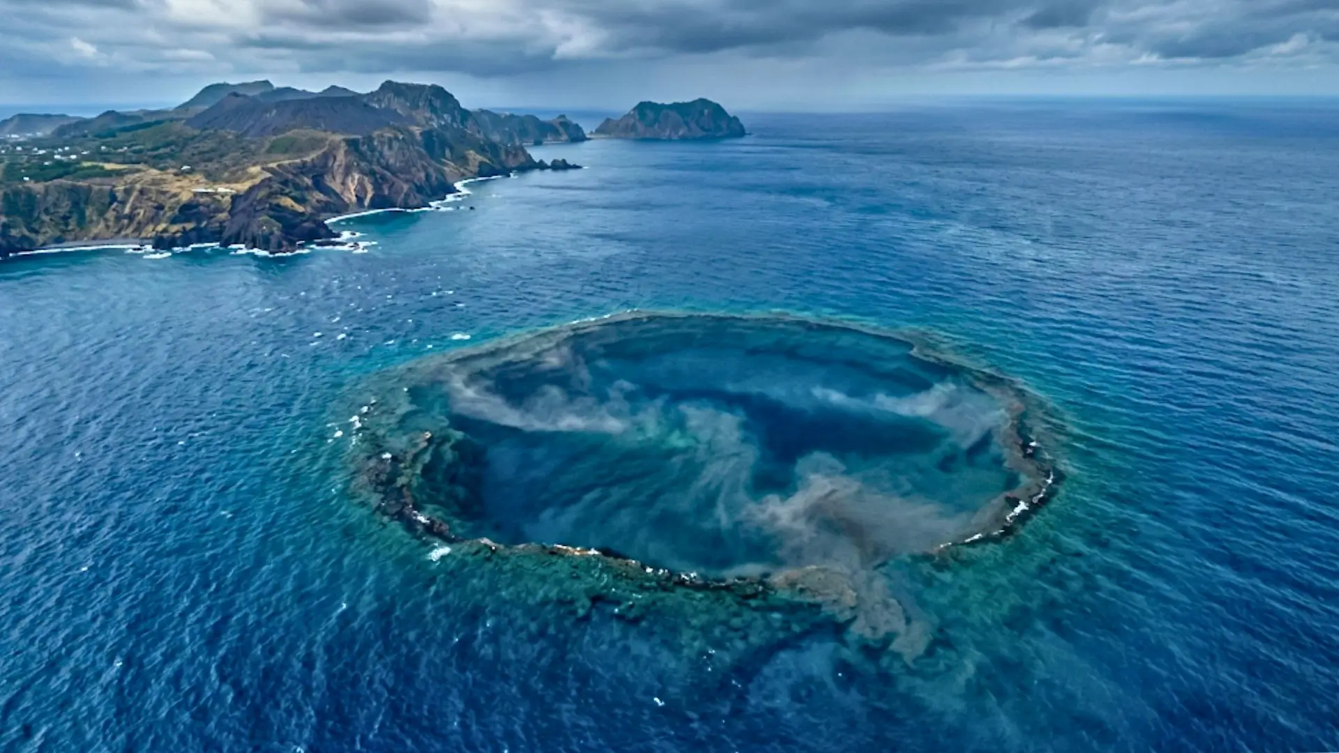 Vue réaliste en plongée aérienne d’une immense caldeira volcanique sous-marine au sud du Japon, visible sous une mer bleu profond près d’îles volcaniques.