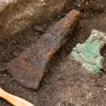 Un artefact en fer très corrodé, en forme de lame de hache primitive, repose à moitié enfoui dans la terre sombre d’une fouille archéologique, à côté d’un fragment de bronze ancien à patine verte.