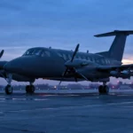 Un petit avion bimoteur à turbopropulseurs stationne sur une piste sombre au crépuscule, avec des drones intercepteurs fixés sous les ailes.