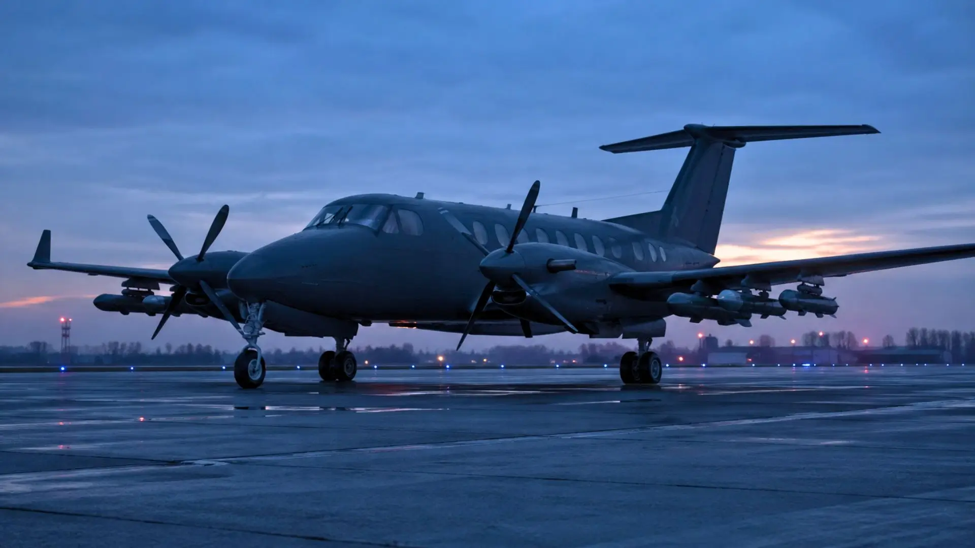 Un petit avion bimoteur à turbopropulseurs stationne sur une piste sombre au crépuscule, avec des drones intercepteurs fixés sous les ailes.