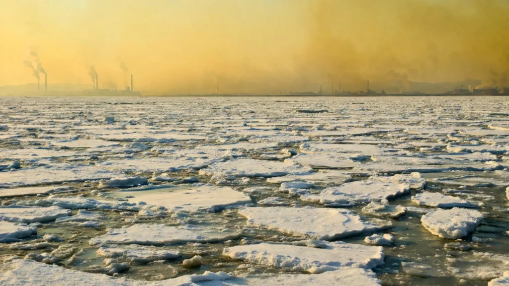Une vaste banquise arctique fracturée s’étend sous un ciel pâle, tandis qu’une brume jaune-brun chargée de pollution recouvre un littoral industriel au loin.