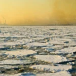 Une vaste banquise arctique fracturée s’étend sous un ciel pâle, tandis qu’une brume jaune-brun chargée de pollution recouvre un littoral industriel au loin.