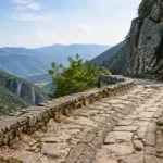 Un sentier pavé en pierres anciennes serpente le long d'une falaise abrupte, surplombant une vaste vallée verdoyante et des montagnes lointaines par une journée ensoleillée.
