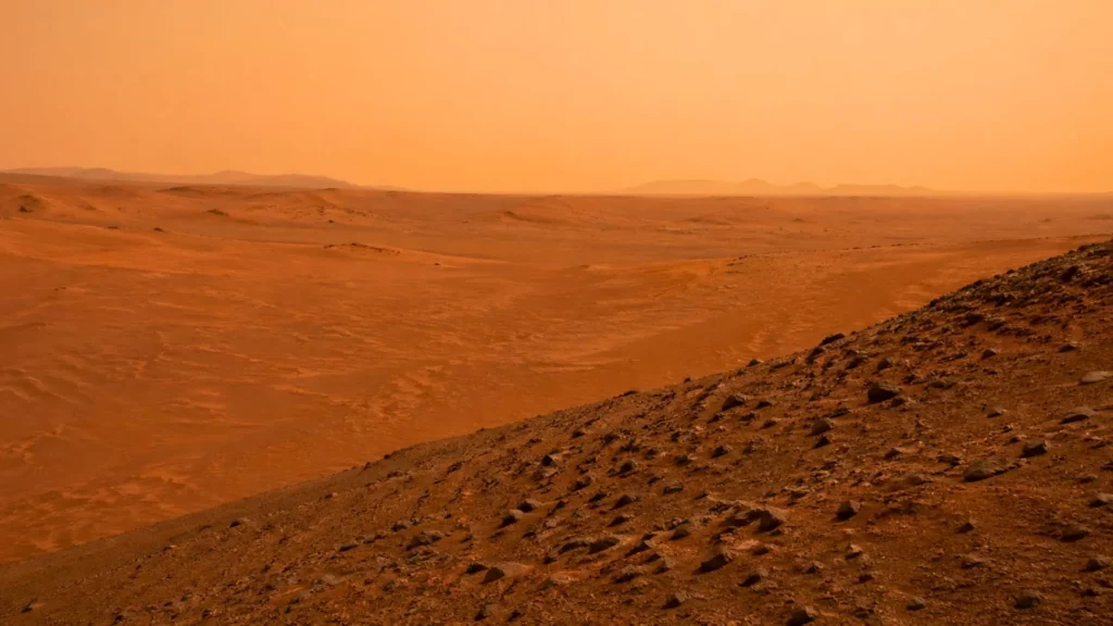 Un vaste désert martien rougeâtre s’étend sous un ciel brumeux, avec des dunes basses, des rides de sable et une pente rocheuse au premier plan.
