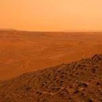 Un vaste désert martien rougeâtre s’étend sous un ciel brumeux, avec des dunes basses, des rides de sable et une pente rocheuse au premier plan.
