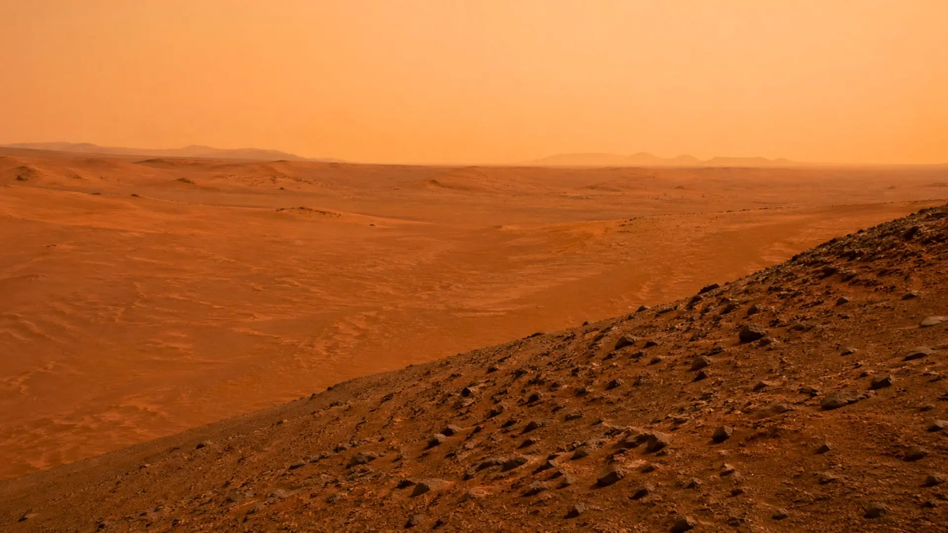Un vaste désert martien rougeâtre s’étend sous un ciel brumeux, avec des dunes basses, des rides de sable et une pente rocheuse au premier plan.
