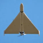 Un drone militaire à aile delta, vu par dessous, vole à basse altitude dans un ciel bleu dégagé.