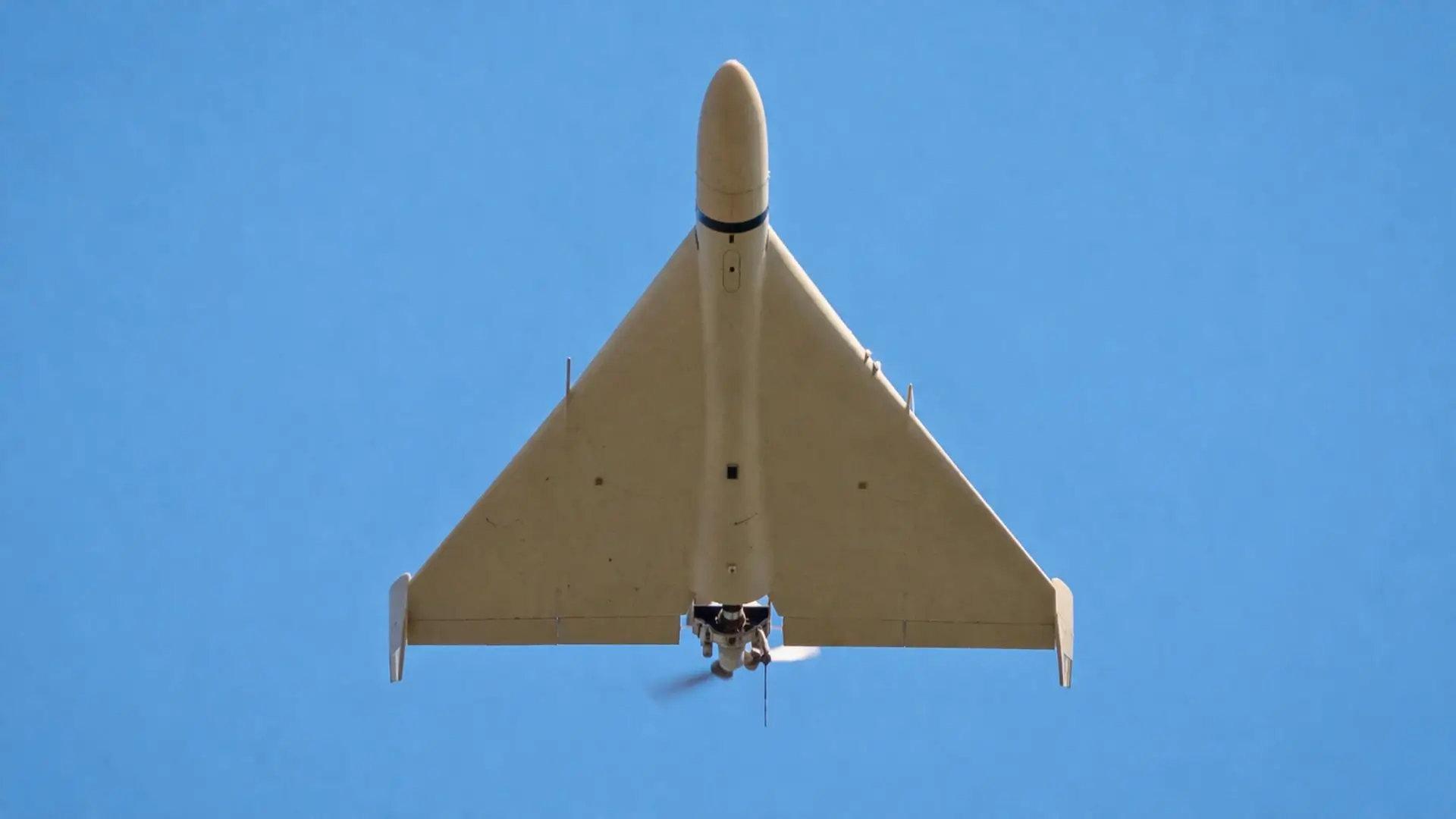 Un drone militaire à aile delta, vu par dessous, vole à basse altitude dans un ciel bleu dégagé.