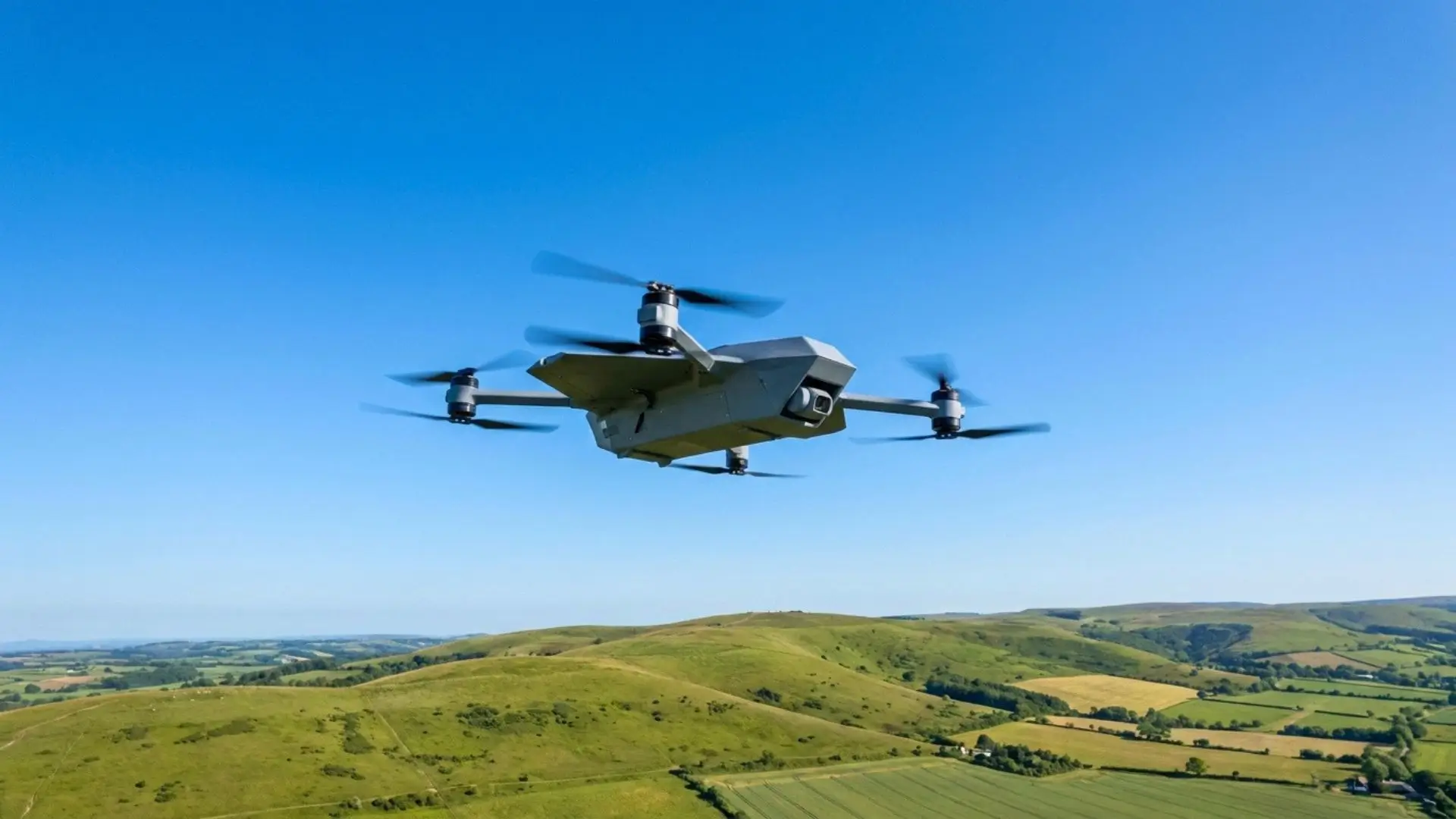 Un drone militaire gris de forme angulaire avec quatre rotors en mouvement, volant dans un ciel bleu clair au-dessus d'un paysage de collines vertes.