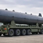 Un drone sous-marin autonome en forme de torpille repose sur un véhicule de transport militaire, sur une vaste esplanade en béton sous un ciel couvert.