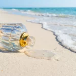 Un flacon de parfum vide en verre ciselé avec des détails dorés est allongé sur le sable blanc, une petite flaque d'eau ou de liquide s'écoulant du goulot vers l'écume de l'océan turquoise.