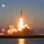 Une immense fusée de mission lunaire décolle au crépuscule depuis une base côtière, avec un épais panache d’échappement au sol et la Lune visible dans le ciel.