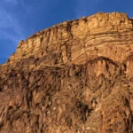 Vue spectaculaire depuis le fond du Grand Canyon vers d’immenses falaises stratifiées, où une nette ligne de discordance sépare des roches très anciennes de couches sédimentaires plus récentes, sous une lumière dorée de fin d’après-midi.